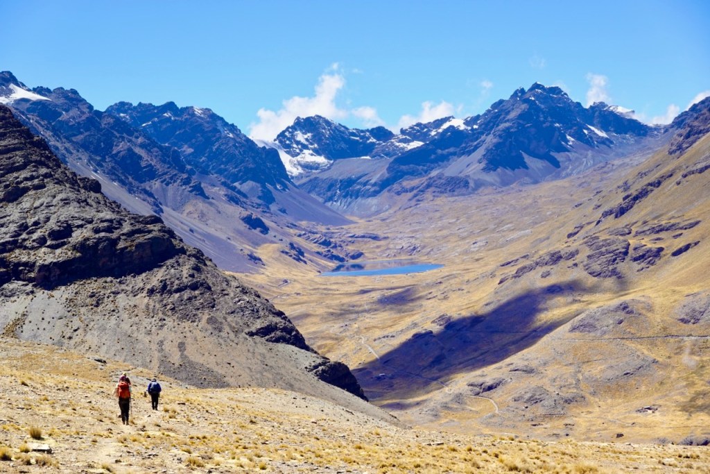 Two hikers walking in a mountainous landscape with rugged terrain and a valley below, featuring a lake surrounded by peaks under a clear blue sky.