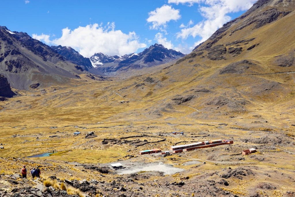 A panoramic view of a mountainous landscape with rugged terrain, featuring snow-capped peaks in the background and a small settlement in the valley below, surrounded by dry, grassy fields.