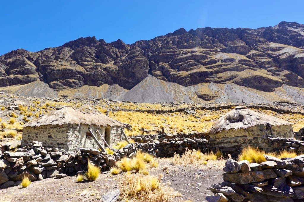 A scenic view of traditional stone huts with thatched roofs in a mountainous region, surrounded by golden grass and rocky hills under a clear blue sky.