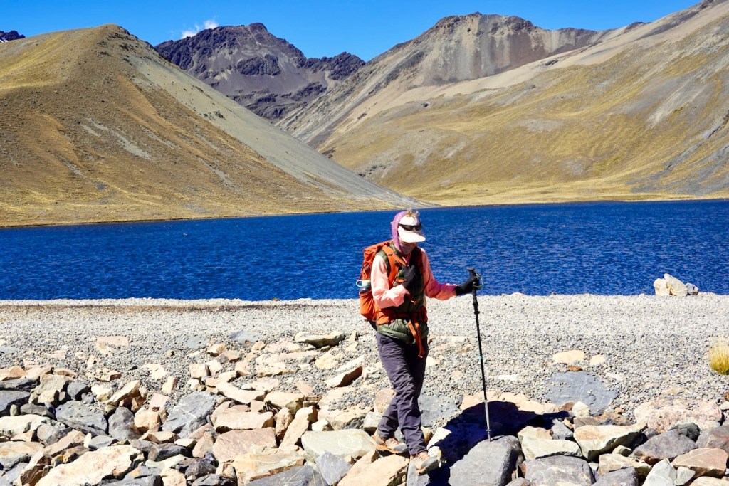 A hiker navigating rocky terrain near a blue lake, surrounded by mountains in a dry landscape.