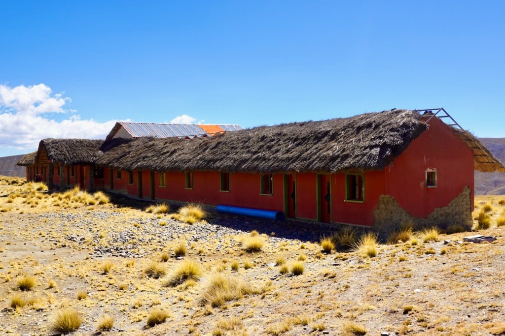 A long, low building with a straw thatched roof and red walls, set against a clear blue sky and surrounded by sparse vegetation on rocky terrain.
