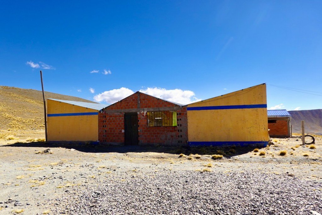 A colorful rural building made of brick and wood with a blue stripe, set against a clear blue sky and mountainous landscape.