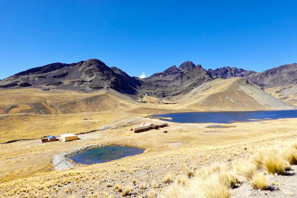 A panoramic view of a mountainous landscape, featuring brown grasslands, rocky slopes, and a large body of water surrounded by two small wooden structures under a clear blue sky.
