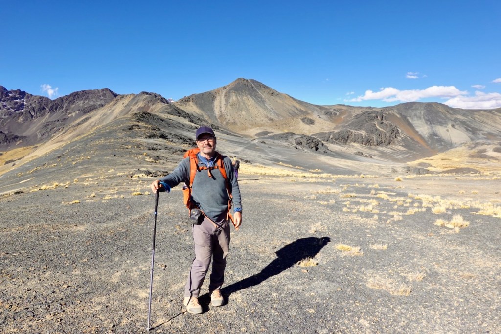 A hiker stands on a rocky landscape with mountains in the background, holding a trekking pole and wearing a backpack.