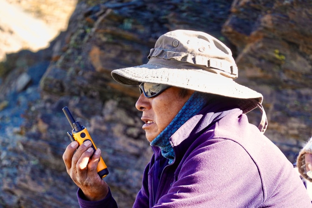 A man wearing a wide-brimmed hat and sunglasses is holding a two-way radio, sitting against a rocky background.