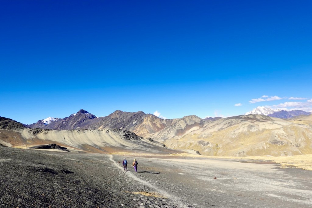 Two hikers walking along a rocky path in a mountainous landscape under a clear blue sky.