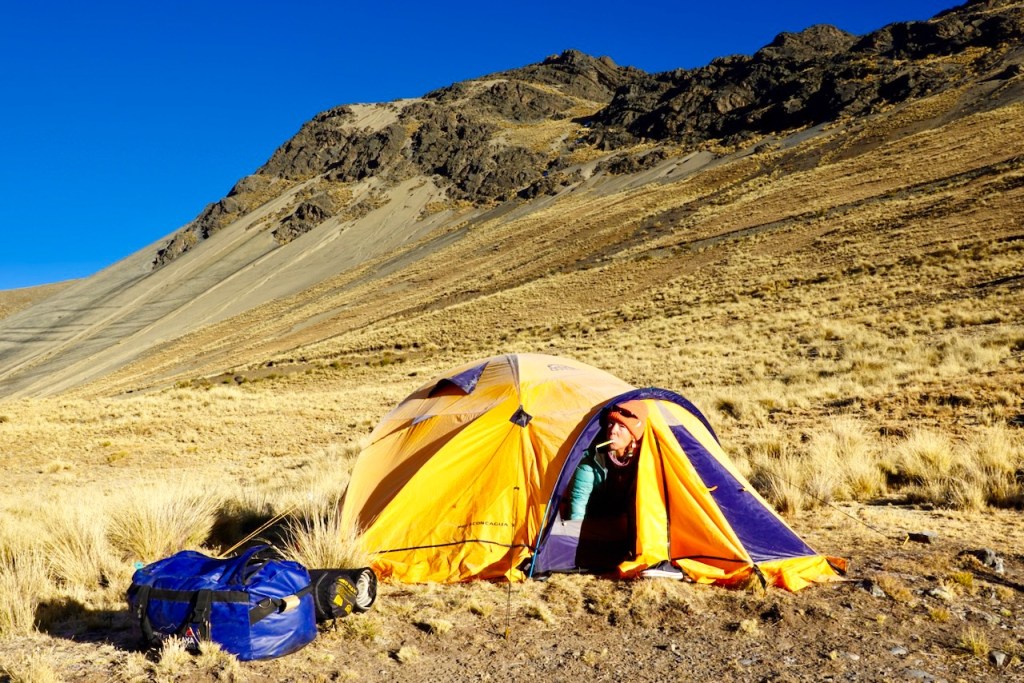 A person emerging from a brightly colored orange and purple tent set against a rugged mountain landscape with dry grass and rocky terrain.