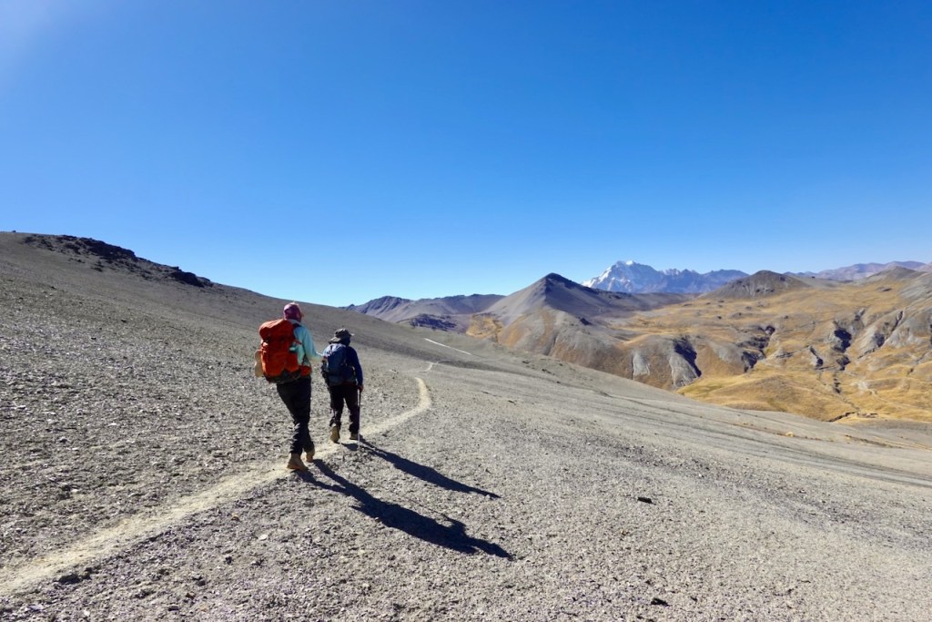 Two hikers walking along a winding dirt trail in a mountainous landscape under a clear blue sky.
