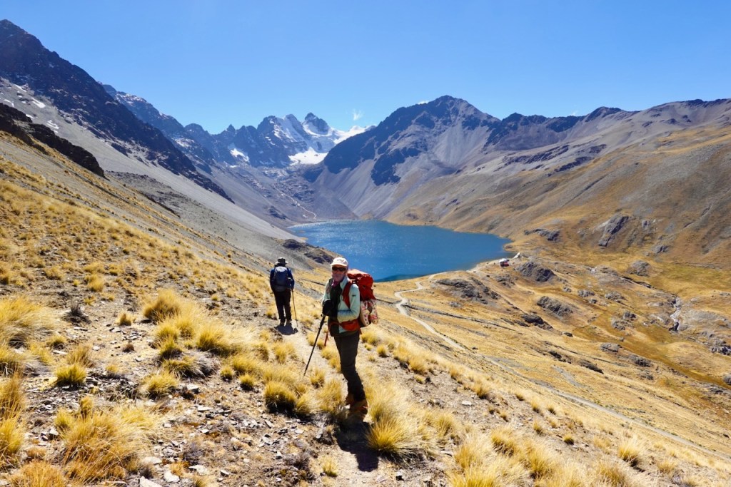 Two hikers trekking on a hillside with golden grass, overlooking a blue lake surrounded by mountains under a clear blue sky.