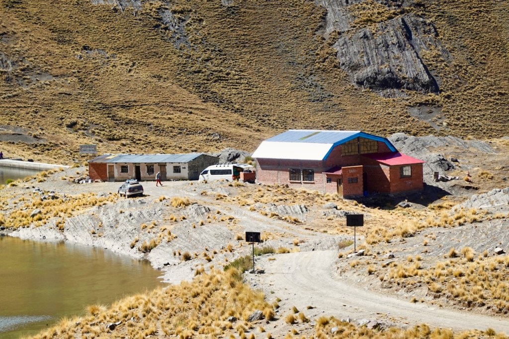 A remote building with two structures, one with a red and blue roof, situated near a pond in a mountainous area with dry grass and rocky terrain.