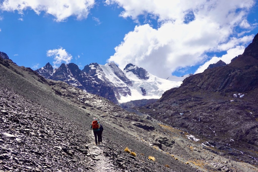 A hiker ascends a rocky path in a mountainous landscape, with snow-capped peaks and a blue sky scattered with clouds in the background.