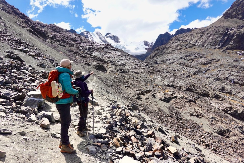 Two hikers on a rocky mountain trail, one pointing towards a glacier in the distance under a blue sky with clouds.