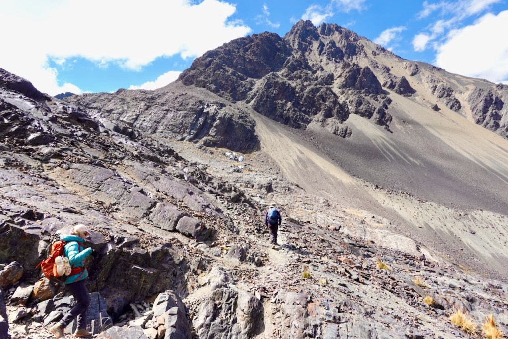 Two hikers trekking on a rocky mountain trail with steep slopes and blue sky in the background.