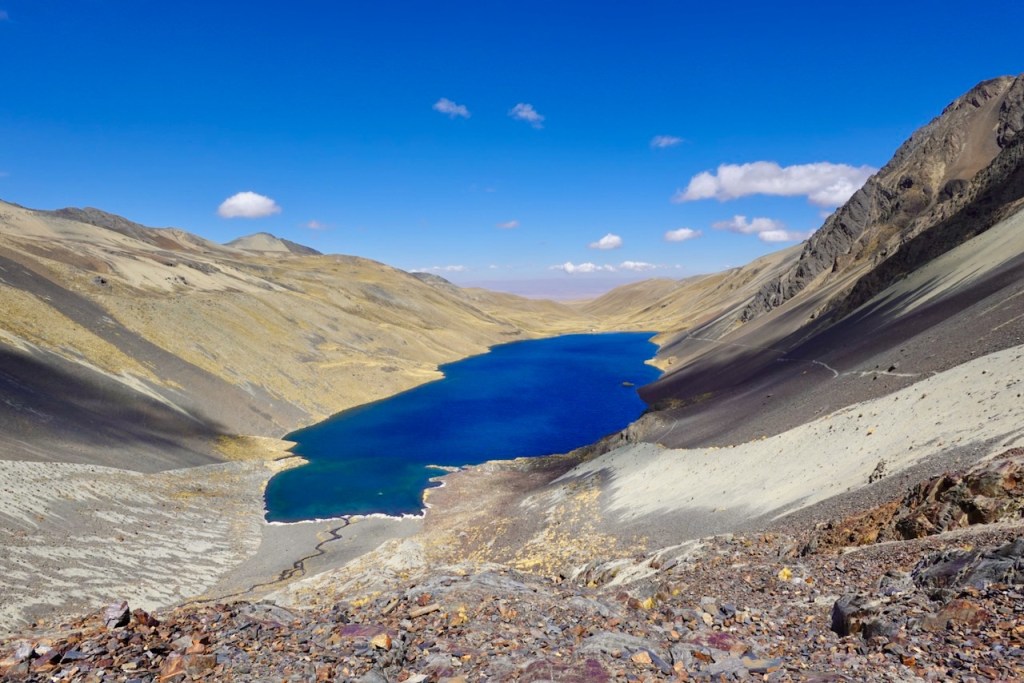 A panoramic view of a vibrant blue lake surrounded by rocky, mountainous terrain under a clear blue sky with a few clouds.