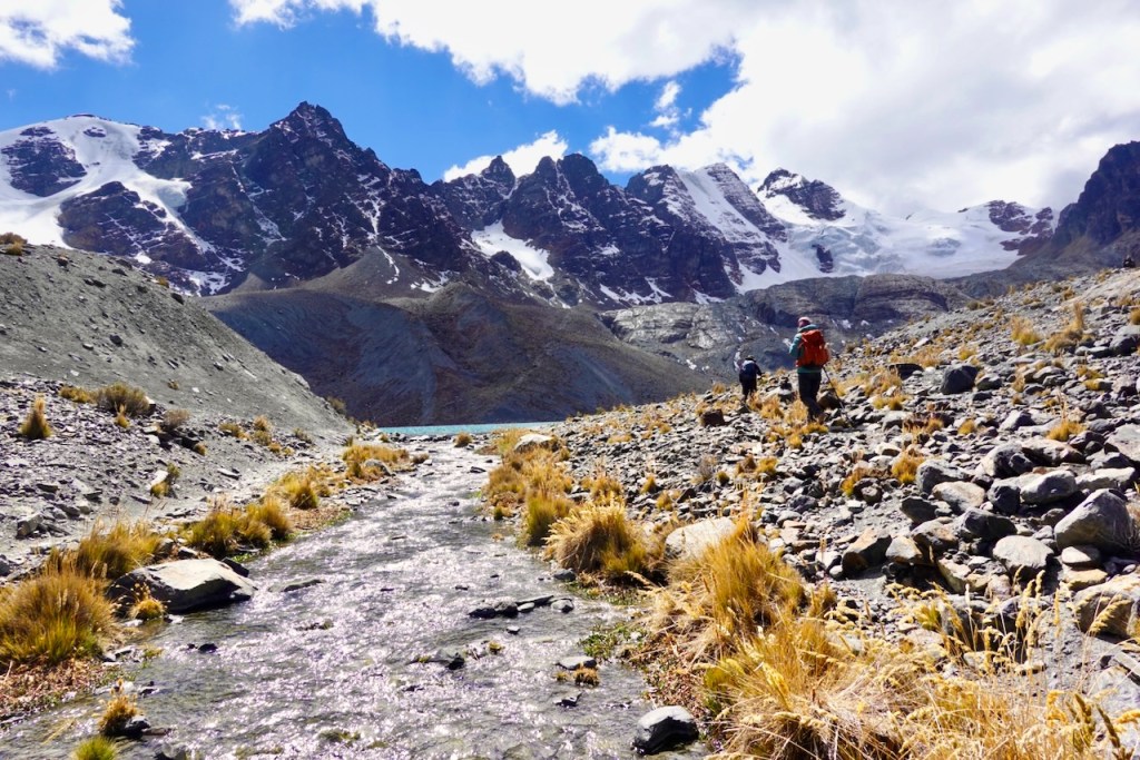 Hikers trekking beside a flowing stream in a mountainous landscape with snow-capped peaks under a bright blue sky.