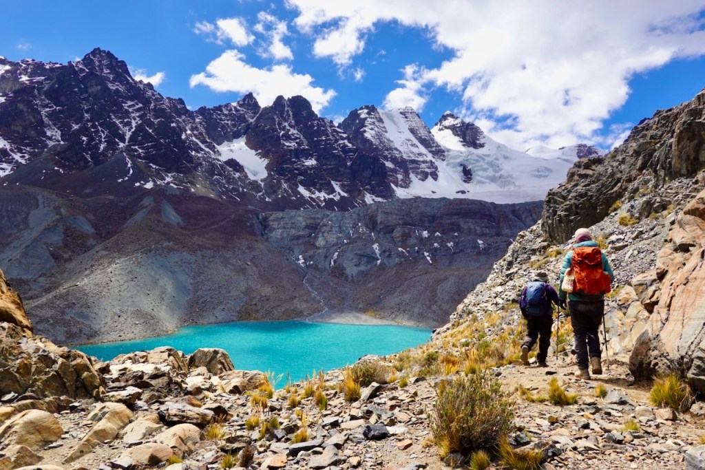 Two hikers walking along a rocky trail near a turquoise lake, surrounded by majestic snow-capped mountains under a partly cloudy sky.