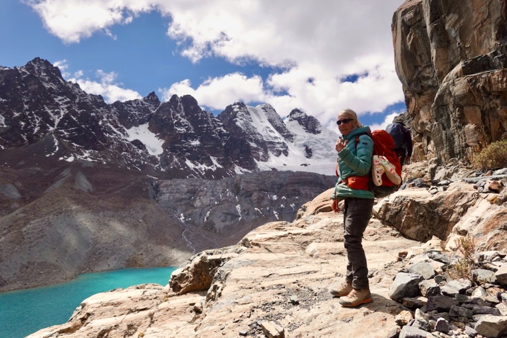 A hiker stands on a rocky trail overlooking a turquoise lake, surrounded by snow-capped mountains and a partly cloudy sky.