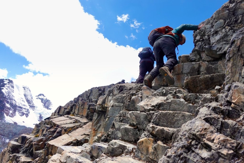 Two hikers climbing a rocky path with snow-capped mountains in the background under a bright blue sky.