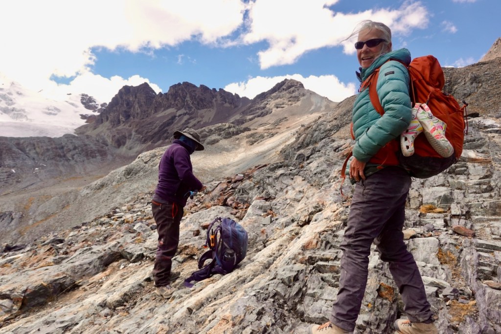 Two hikers on a rocky mountain terrain, one standing and smiling with a backpack, and the other bent down near a backpack, with mountains and blue sky in the background.