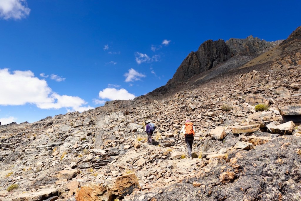 Two hikers ascending a rocky mountain trail under a blue sky with scattered clouds.