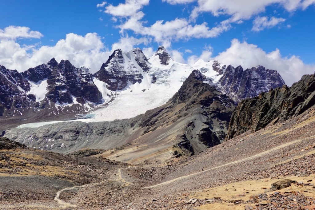 A panoramic view of rugged mountains with snow-capped peaks under a blue sky filled with clouds, showcasing rocky terrain and a winding path in the foreground.