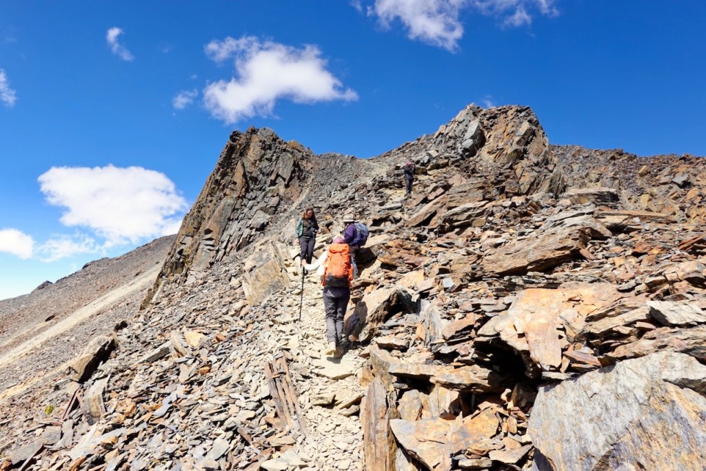 Hikers ascending a rocky mountain trail under a bright blue sky with fluffy clouds.