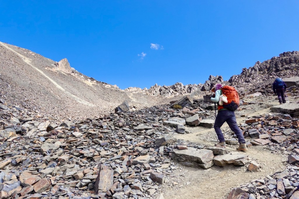 Hikers trekking on a rocky mountain trail under a clear blue sky.