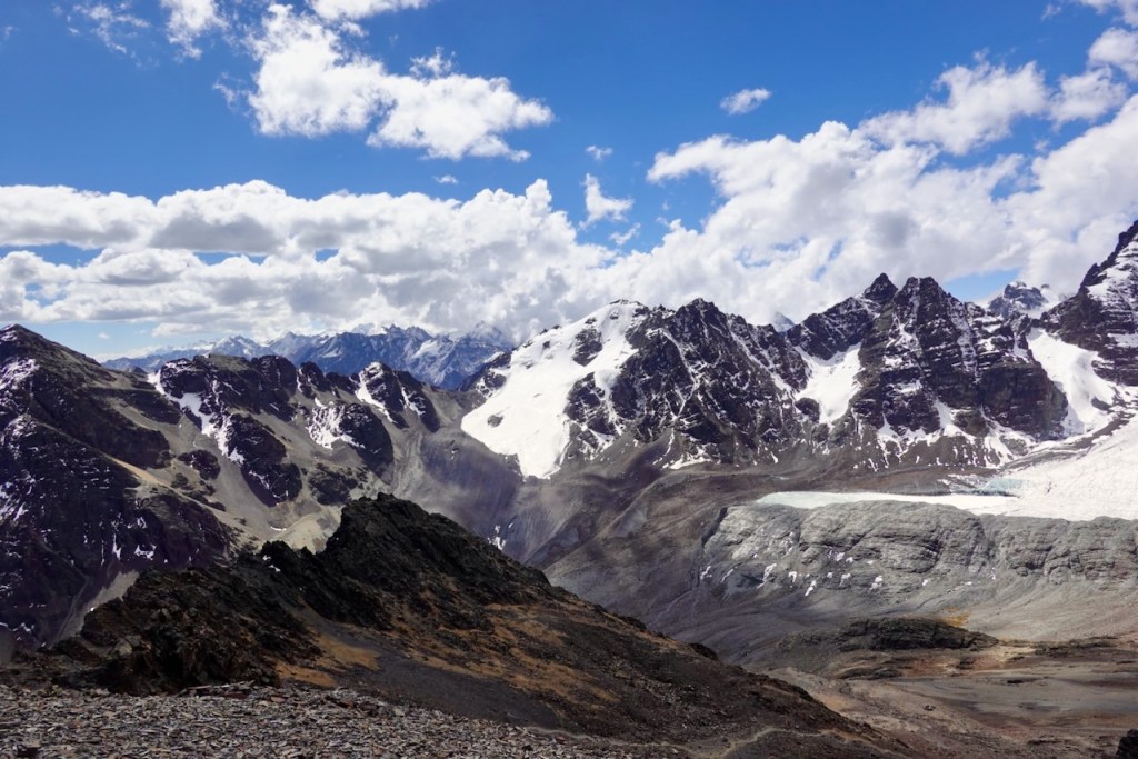 A panoramic view of snow-capped mountains under a partly cloudy blue sky, showcasing rocky terrain and glaciers.