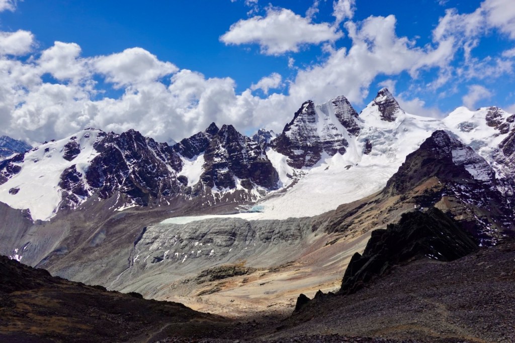 A panoramic view of majestic snow-capped mountains under a partly cloudy sky, showcasing rugged terrain and a glacier in the foreground.
