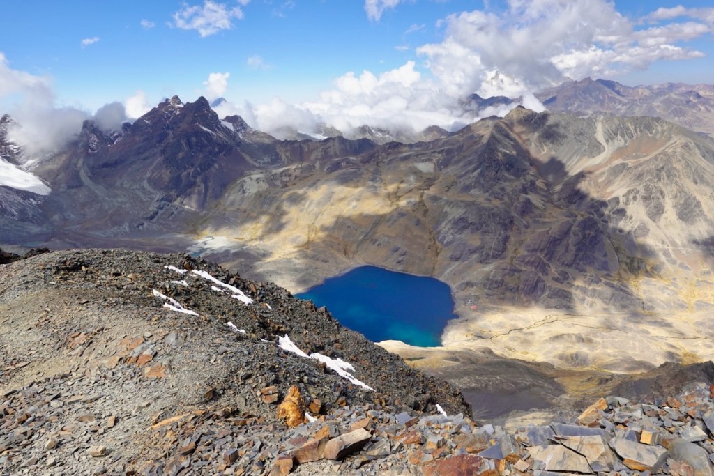 A panoramic view of a mountain landscape featuring jagged peaks and a deep blue lake surrounded by rocky terrain and clouds in the sky.
