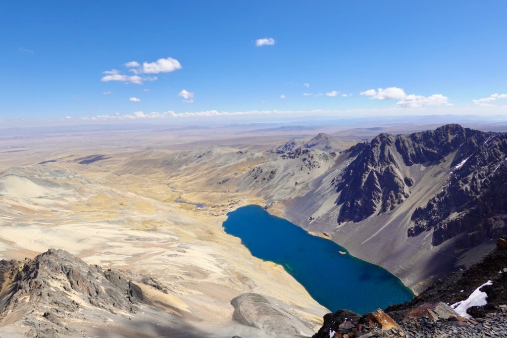 A panoramic view of a mountainous landscape featuring a bright blue lake surrounded by rocky terrain and sparse vegetation under a clear sky with a few clouds.