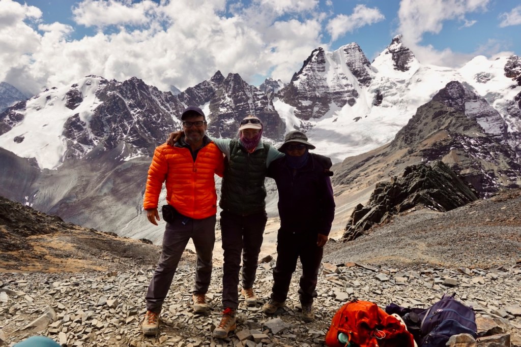 Three hikers posing on a mountain ridge with snow-capped peaks in the background.