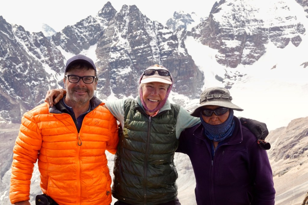 Three climbers posing together in the mountains, with snow-capped peaks in the background. They are wearing brightly colored jackets and sunglasses, smiling at the camera.