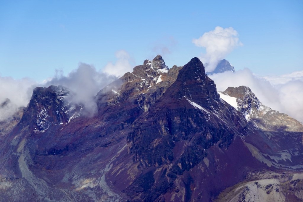 A dramatic mountain range with jagged peaks, partially covered in snow and surrounded by clouds, under a clear blue sky.