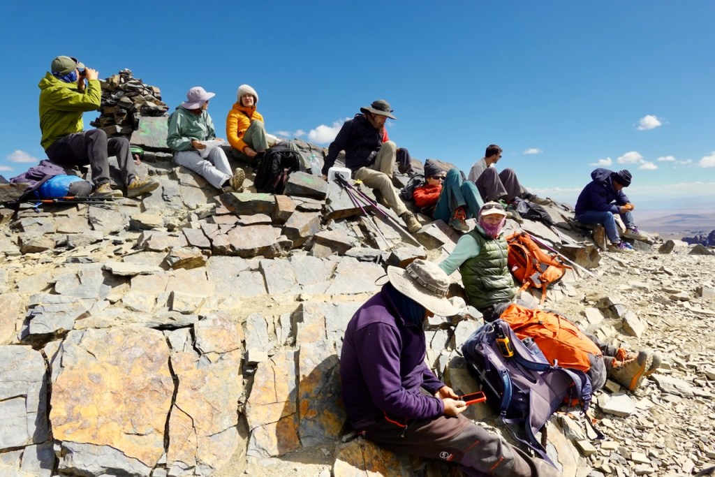 A group of hikers resting on rocky terrain at a mountain peak, with clear blue skies and scattered clouds in the background.
