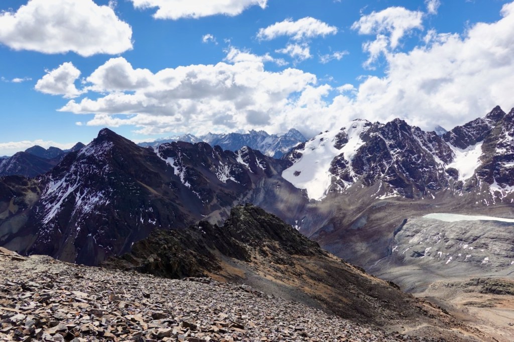 Scenic view from the top of a mountain range featuring snow-capped peaks, rocky terrain, and a blue sky with scattered clouds.