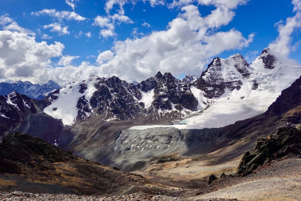 A panoramic view of a snow-capped mountain range under a blue sky with fluffy white clouds, showcasing rocky terrain and a glacier in the foreground.
