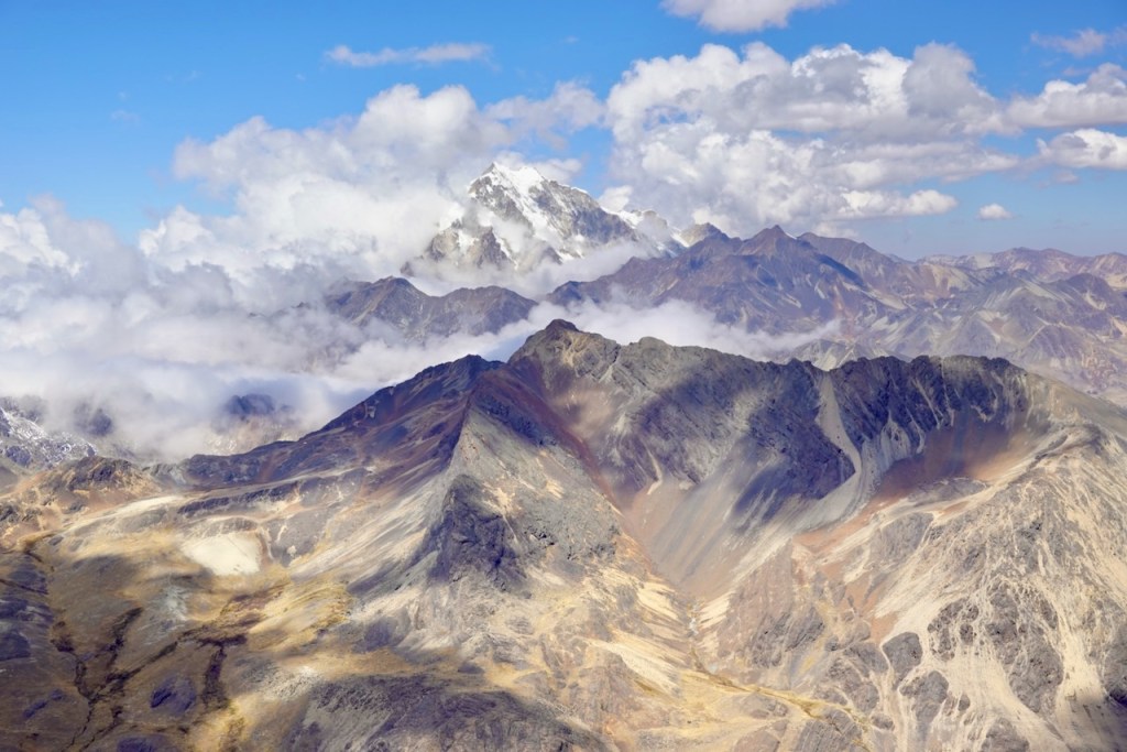 Aerial view of majestic mountains with peaks partially covered in clouds and a clear blue sky, showcasing rugged terrain and varied colors of the landscape.