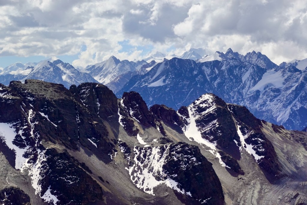 A panoramic view of snow-capped mountains under a partly cloudy sky, showcasing rugged peaks and rocky terrain.