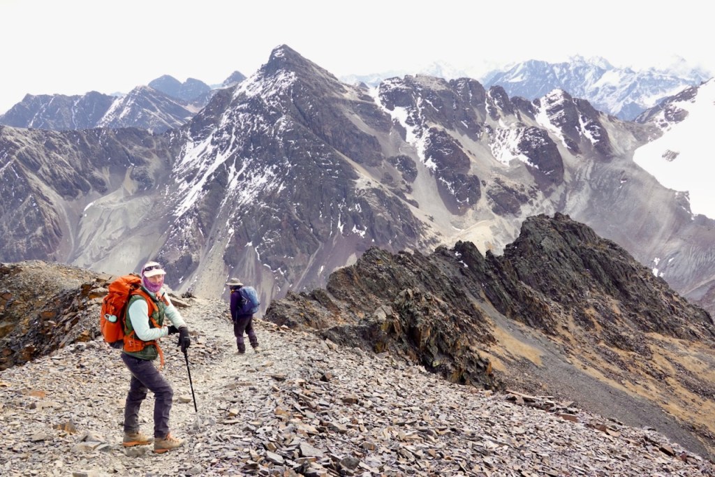 Two hikers navigating a rocky mountain trail with snow-capped peaks in the background.