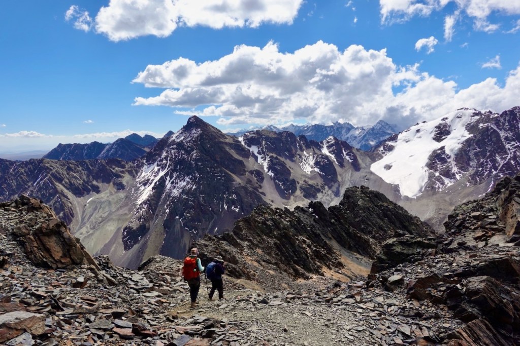 Two hikers walking along a rocky trail with snow-capped mountains in the background under a blue sky with fluffy clouds.