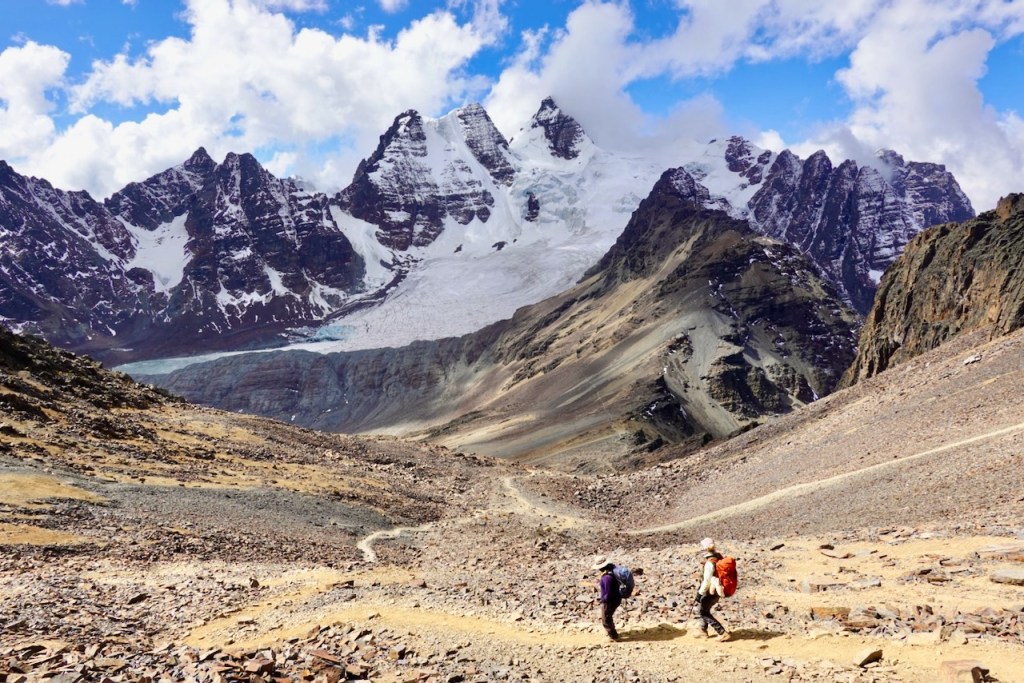 Two hikers navigating a rocky trail in a mountainous landscape with snow-capped peaks and a glacier in the background.