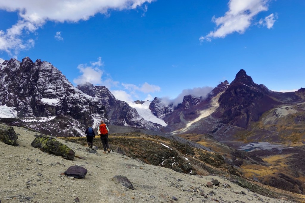 Two hikers walking on a trail in a mountainous landscape with snow-capped peaks under a blue sky with scattered clouds.