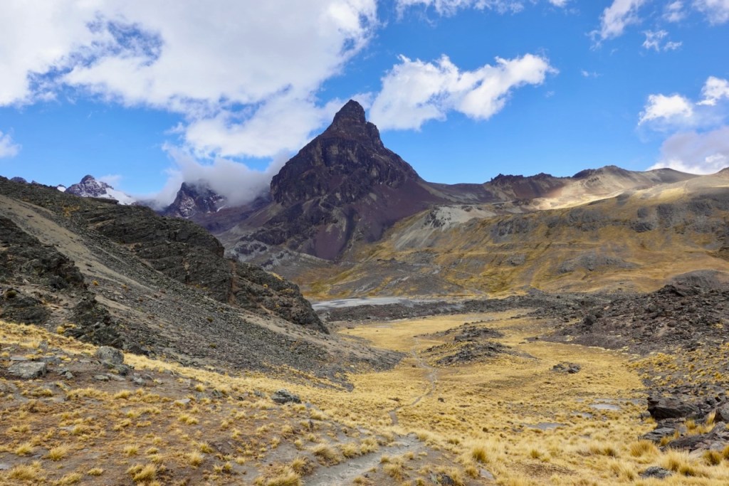 A scenic view of a mountainous landscape featuring a prominent peak under a blue sky with scattered clouds, surrounded by rocky terrain and sparse vegetation.