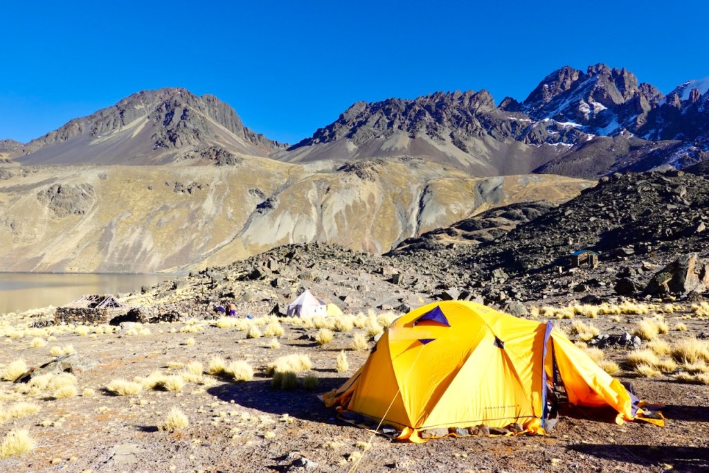 A yellow tent set up on rocky terrain by a lake, surrounded by brown mountains and sparse grass under a clear blue sky.