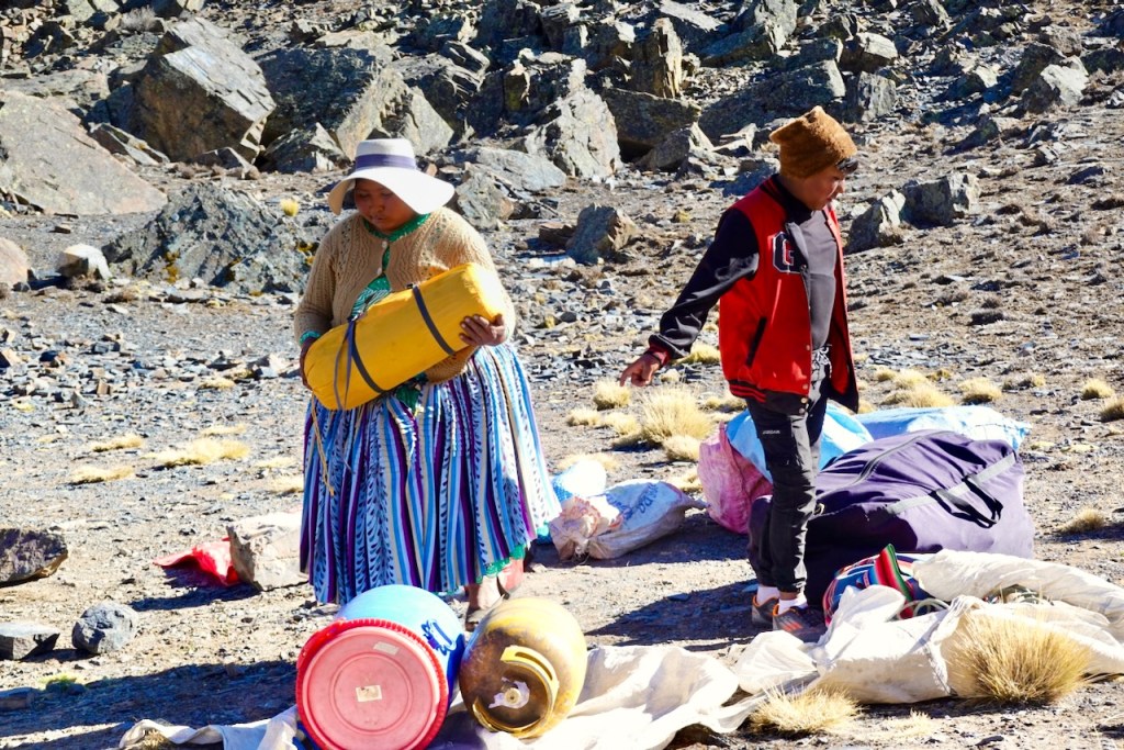 A woman in traditional clothing carrying a large yellow container while a boy walks nearby on rocky terrain.