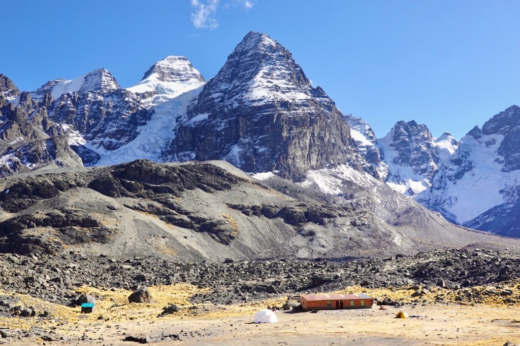 A vast mountain landscape featuring snow-capped peaks, rocky terrain, and a small building surrounded by tents, under a clear blue sky.