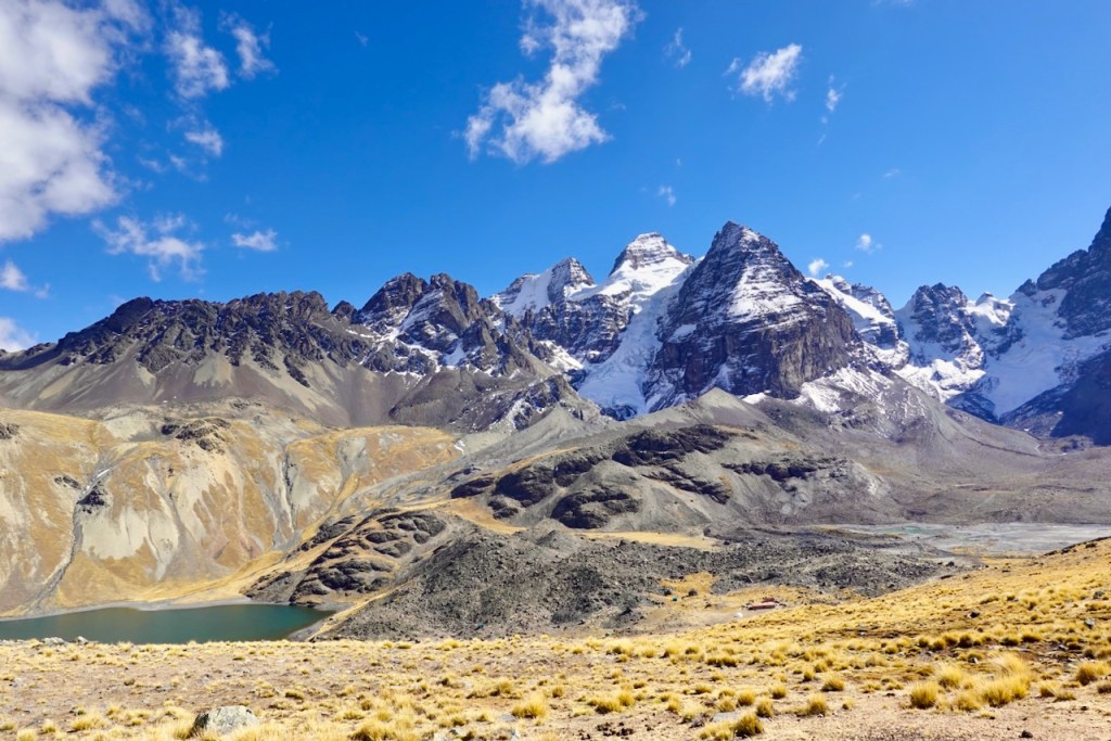 A panoramic view of towering snow-capped mountains under a blue sky, with a tranquil lake and rocky terrain in the foreground.