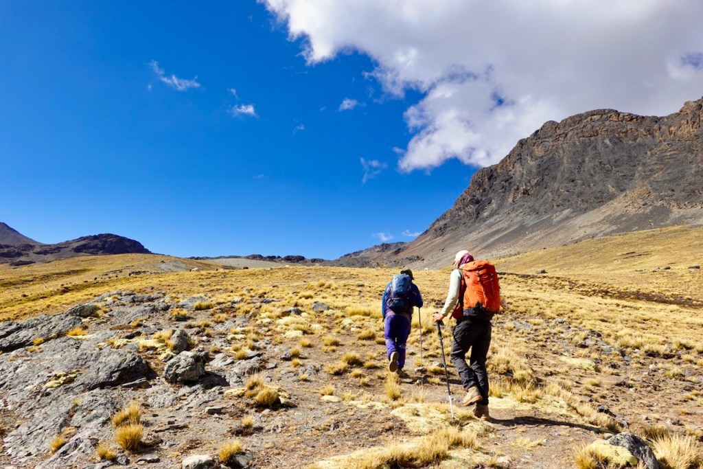 Two hikers ascending a rocky path in a mountainous landscape under a bright blue sky with scattered clouds.