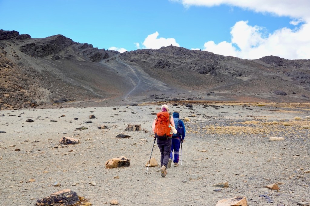 Two hikers walking on a rocky terrain with mountains in the background under a partly cloudy sky.